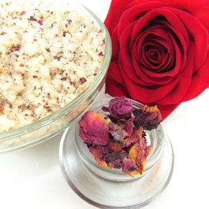 Explore a tub of exfoliating cane sugar scrub, infused with fragrant red rose petals. The scrub sits in a glass bowl next to a vibrant, fully bloomed red rose. Dried rose petals adorn a smaller glass bowl in the foreground against a white backdrop.