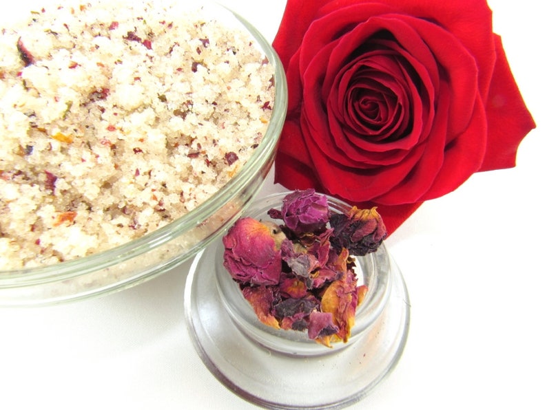 Explore a tub of exfoliating cane sugar scrub, infused with fragrant red rose petals. The scrub sits in a glass bowl next to a vibrant, fully bloomed red rose. Dried rose petals adorn a smaller glass bowl in the foreground against a white backdrop.