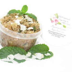 A 2 oz tub of cooling peppermint and coconut cane sugar scrub offers a refreshing and hydrating experience for your skin. The sugar scrub is centered, near bright green sprigs of peppermint and coconut flakes are scattered throughout the arrangement.