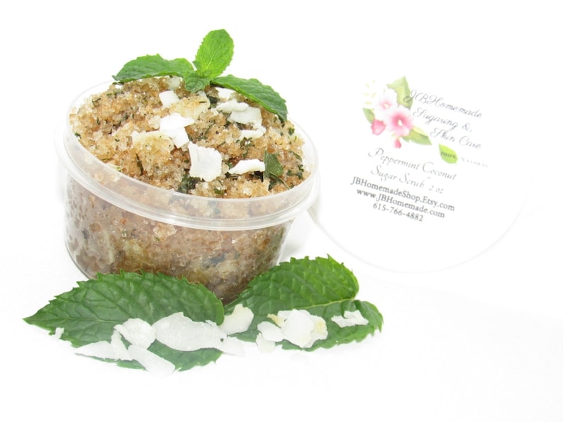 A 2 oz tub of cooling peppermint and coconut cane sugar scrub offers a refreshing and hydrating experience for your skin. The sugar scrub is centered, near bright green sprigs of peppermint and coconut flakes are scattered throughout the arrangement.