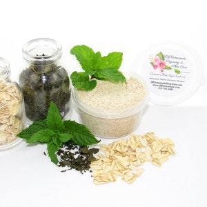 A 2 oz tub of gentle exfoliating dry body scrub, ideal for sensitive skin, featuring colloidal oatmeal, fresh and dried mint, and brown sugar. Accompanying the scrub are oats and dried mint spilled on a white table by sprigs of green fresh mint.