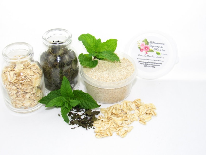 A 2 oz tub of gentle exfoliating dry body scrub, ideal for sensitive skin, featuring colloidal oatmeal, fresh and dried mint, and brown sugar. Accompanying the scrub are oats and dried mint spilled on a white table by sprigs of green fresh mint.