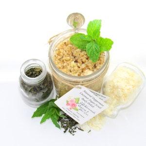 An 8 oz jar of peppermint and coconut cane sugar scrub offers a refreshing and hydrating experience for your skin. The sugar scrub is centered, near bright green sprigs of peppermint, dried mint and coconut flakes are scattered in the arrangement.