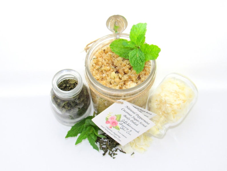 An 8 oz jar of peppermint and coconut cane sugar scrub offers a refreshing and hydrating experience for your skin. The sugar scrub is centered, near bright green sprigs of peppermint, dried mint and coconut flakes are scattered in the arrangement.