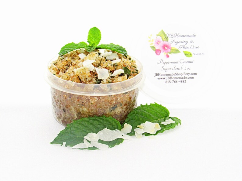 A 2 oz tub of cooling peppermint and coconut cane sugar scrub offers a refreshing and hydrating experience for your skin. The sugar scrub is centered, near bright green sprigs of peppermint and coconut flakes are scattered throughout the arrangement.
