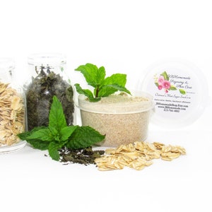 May include: A clear glass jar filled with rolled oats, a clear glass jar filled with green tea leaves, a sprig of fresh mint, and a clear plastic container filled with a light brown sugar scrub. The container has a label that reads "Homemade Regimen & Skin Care" and "Oatmeal Mint Sugar Scrub".