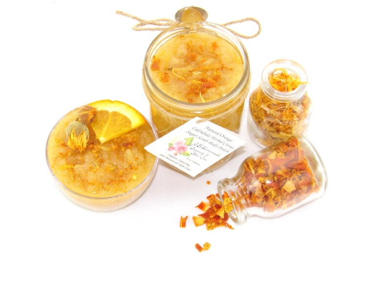 An 8 oz jar of citrus sugar scrub, with orange & calendula, for a radiant glow. The jar is centered by 2 small jars, 1 with orange zest, 1 with calendula spilling on a white table. A bowl of sugar scrub, with calendula petals and an orange slice.