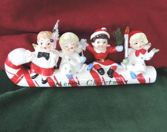 Könnte beinhalten: Eine weihnachtliche Keramikfigur mit vier Kindern, die auf einer großen weißen Zuckerstange mit roten Streifen sitzen. Auf der Basis steht der Text Merry Christmas. Die Kinder tragen festliche Kleidung und halten kleine Accessoires.