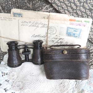 May include: Antique opera glasses with a dark brown leather case and a bundle of vintage letters tied with twine. The opera glasses are black and brown, and the letters have postage stamps.