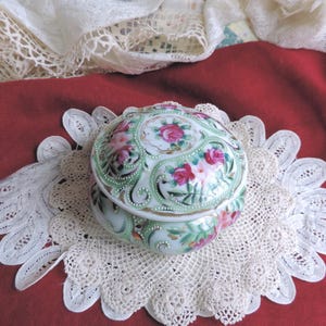 May include: A round porcelain trinket box featuring a hand-painted floral motif with pink roses and green foliage. The lid and base are decorated with raised white enamel dot accents and gold trim, showcasing a scalloped edge design throughout.