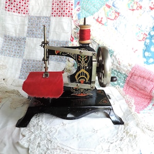 May include: A vintage black cast iron sewing machine featuring decorative floral patterns in red, yellow, and green. The machine has a silver hand crank wheel, a vertical spool pin holding a red thread spool, and a piece of red fabric under the needle.