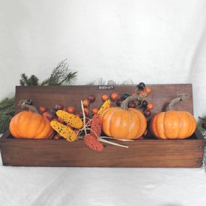 May include: A rectangular dark brown wooden box filled with three orange pumpkins, clusters of small red and orange berries, and yellow textured corn-like decorative pieces, accented by green pine branches on the sides.