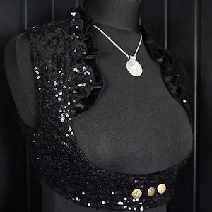 May include: A black sequined and velvet cropped vest with a ruffled neckline and three gold buttons. A silver necklace with a round pendant hangs from the vest.