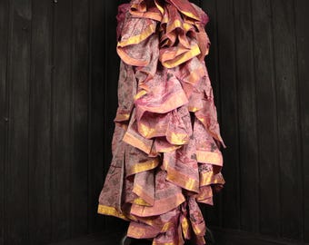 May include: A layered, ruffled skirt in shades of pink and mauve, with gold trim. The skirt is displayed on a black mannequin against a dark wood background. The fabric has a subtle pattern.