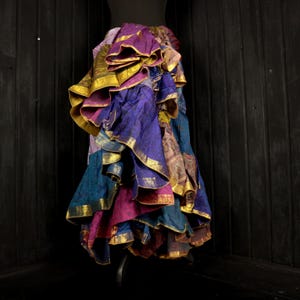 Könnte beinhalten: Ein mehrschichtiger Rock in Lila-, Türkis-, Rosa- und Goldtönen. Der Rock hat goldfarbene Verzierungen und ein einzigartiges Design. Das Kleidungsstück wird auf einer Schaufensterpuppe präsentiert. Das Design des Rocks ist auffällig und die Farben sind lebendig.