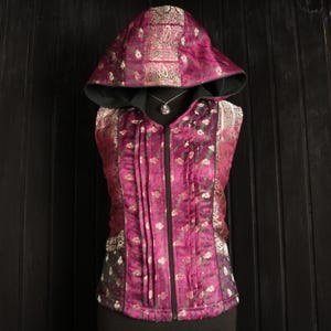 May include: A sleeveless hooded vest with a zip closure. The vest is made of a pink fabric with a gold and green floral pattern. The hood is lined with black fabric.