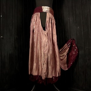 Puede incluir: Una falda larga y fluida en color rosa empolvado, con una cinturilla ancha y adornada. La falda tiene una abertura frontal y un dobladillo burdeos en contraste. Un panel burdeos a juego con detalles plateados cae a un lado.