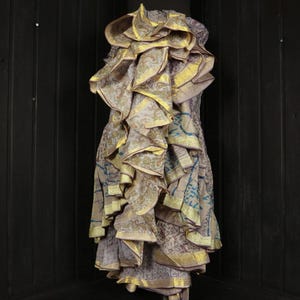 May include: A unique, layered dress with a ruffled neckline and skirt. The dress features a mix of patterned fabrics in shades of beige, gold, and blue. The ruffles are trimmed with gold accents, creating a textured and elegant look.