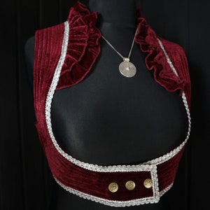 Può includere: Un gilet di velluto rosso con un bordo in pizzo bianco e tre bottoni dorati. Il gilet ha un colletto a volant e una collana con un ciondolo argentato di forma circolare.