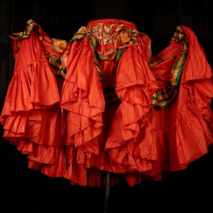 May include: A vibrant, ruffled skirt in a bright orange hue. The skirt features multiple layers of fabric, creating a full, flowing silhouette. Green and gold trim accents the waist and edges, adding a touch of contrast. The skirt appears to be made of a lightweight material.