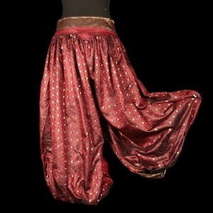 Peut inclure: Pantalon sarouel rouge foncé en tissu à motifs dorés métallisés avec un léger éclat. Le vêtement présente une large ceinture texturée de couleur bronze et des poignets assortis aux chevilles, créant une silhouette volumineuse.