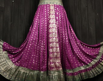 May include: A flowing, floor-length skirt in a deep purple hue, adorned with gold polka dots and intricate floral patterns. The skirt features a wide waistband with green and gold trim, and a ruffled hemline. The fabric drapes elegantly, creating a voluminous silhouette.