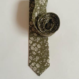 May include: Olive green necktie with a white floral pattern. The tie is partially rolled up, showcasing the fabric's design. The tie's fabric has a matte finish, and the floral pattern is evenly distributed across the surface.