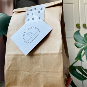 May include: A brown paper bag sealed with polka dot tape and a tag that reads "THE LITTLE GREYHOUND handmade in England happy mail!" The bag is held in front of a leafy green plant.