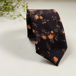 May include: A dark navy tie with a repeating floral pattern of orange and brown flowers and leaves. The tie is folded over on a white surface, with a hint of greenery in the background.