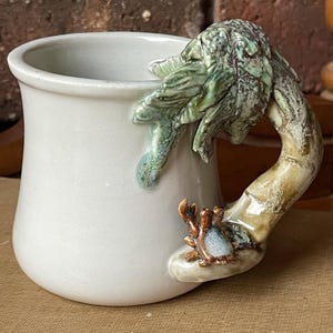 May include: White ceramic mug with a unique handle shaped like a branch with green leaves and a small crab. The mug has a smooth, glossy finish and a slightly flared rim. The handle is detailed with textured, earthy tones.
