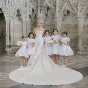 Può includere: Una sposa in un abito da sposa bianco con un lungo strascico sta con cinque damigelle d'onore in abiti bianchi. Le ragazze tengono mazzi di fiori bianchi. Il gruppo è in piedi in una grande stanza ornata con porte ad arco.