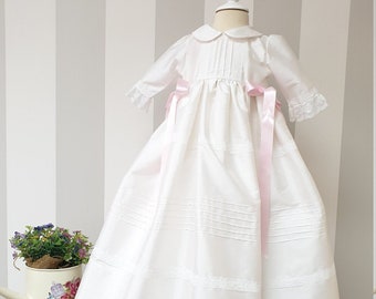 May include: A white christening gown with a pink satin sash and lace trim. The gown has a long skirt and a fitted bodice with a peter pan collar.