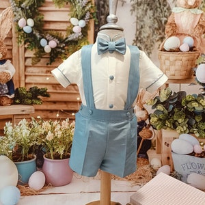 May include: A child's light blue shorts set, comprising a white shirt with blue trim, matching shorts with braces, and a bow tie. The outfit is displayed on a mannequin with beige shoes. The background features Easter decorations.