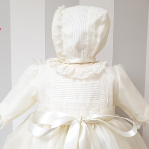 Peut inclure: Une robe de bapt&ecirc;me blanche avec une bordure en dentelle et un bonnet assorti. La robe est orn&eacute;e d'un ruban de satin nou&eacute; &agrave; la taille.