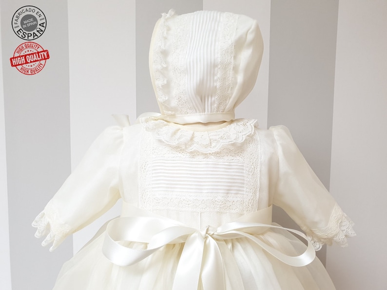 Peut inclure: Une robe de bapt&ecirc;me blanche avec une bordure en dentelle et un bonnet assorti. La robe est orn&eacute;e d'un ruban de satin nou&eacute; &agrave; la taille.