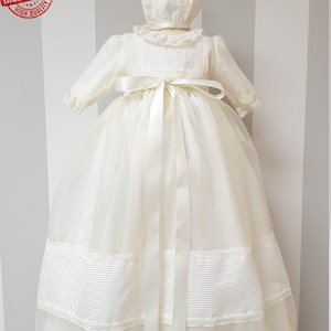 Peut inclure: Une robe de bapt&ecirc;me blanche avec une bordure en dentelle et un ruban de satin. La robe est assortie &agrave; un bonnet avec une bordure en dentelle.