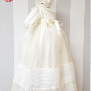 Peut inclure: Une robe de bapt&ecirc;me blanche avec une bordure en dentelle et une ceinture en satin. La robe est assortie &agrave; un bonnet avec une bordure en dentelle.
