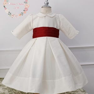 Puede incluir: Vestido blanco con fajín rojo, exhibido en un maniquí. El vestido tiene cuello redondo y mangas largas. La falda es plisada y acampanada. El vestido está diseñado para niños. La marca "Maria Platerie Kids" es visible en el fondo.