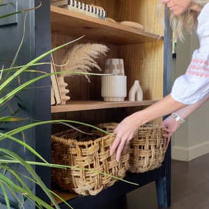 K&ouml;nnte beinhalten: Ein schwarz lackierter Holzschrank mit Regalen. Die Regale sind mit Dekorationsartikeln gef&uuml;llt, darunter eine Vase, ein Buch und ein Weidenkorb. Der Schrank steht auf einem dunklen Holzboden.