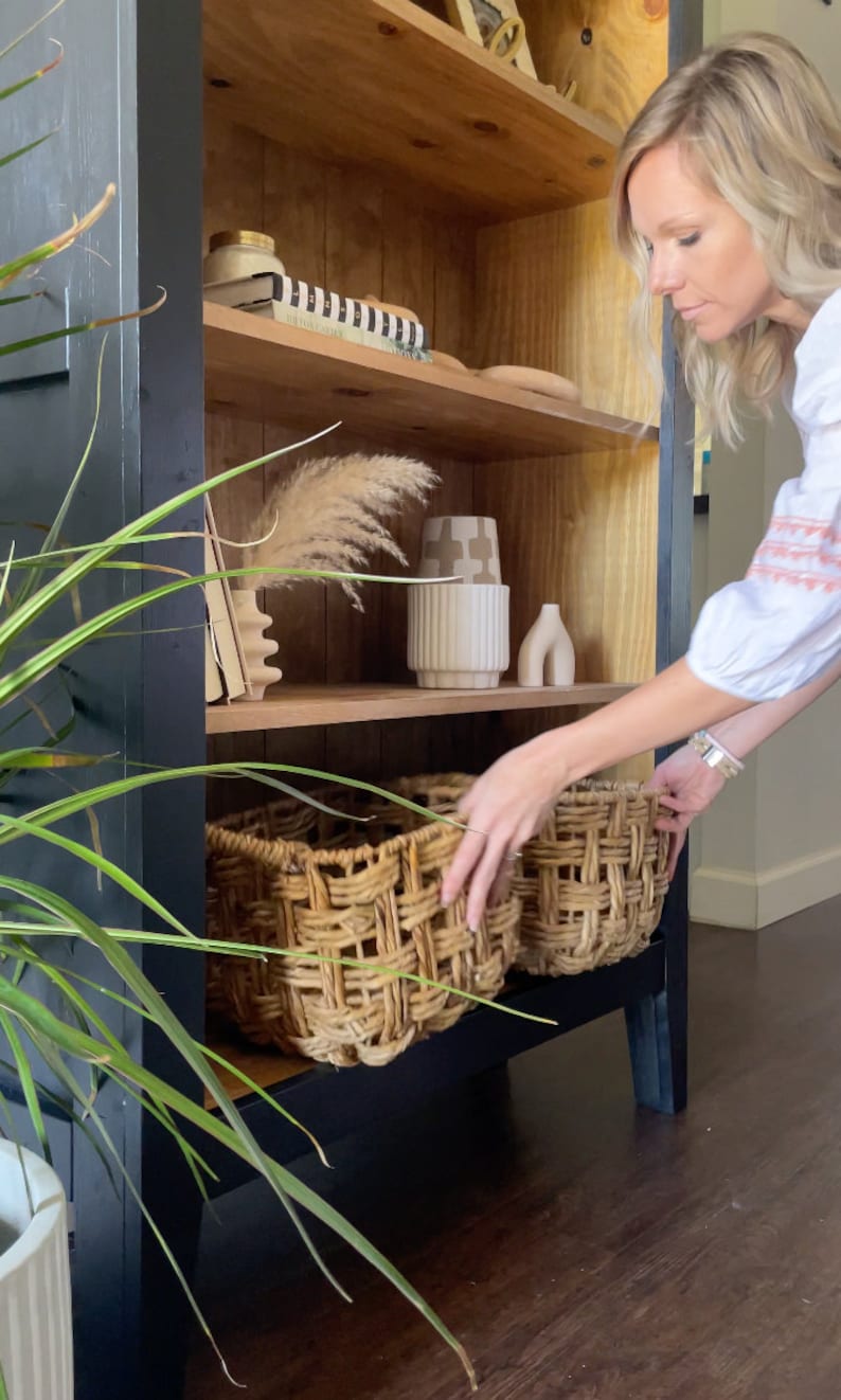 K&ouml;nnte beinhalten: Ein schwarz lackierter Holzschrank mit Regalen. Die Regale sind mit Dekorationsartikeln gef&uuml;llt, darunter eine Vase, ein Buch und ein Weidenkorb. Der Schrank steht auf einem dunklen Holzboden.