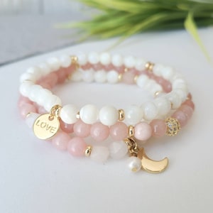 Peut inclure: Deux bracelets superposés avec des perles roses et blanches. Le bracelet du haut a un charme en or avec le mot "LOVE" dessus. Le bracelet du bas a un charme en or en forme de croissant de lune avec une petite perle.