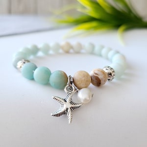 May include: A light blue and beige beaded bracelet with a silver starfish charm and a pearl. The bracelet is on a white surface.