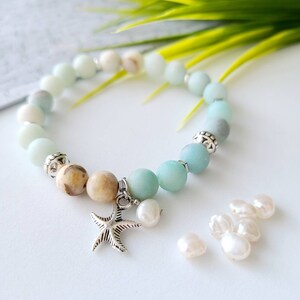 May include: A light blue and brown beaded bracelet with a silver starfish charm. The bracelet is made with natural stones and has a gradient of colors. The bracelet is accompanied by several white pearl beads.