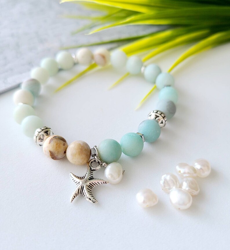May include: A light blue and brown beaded bracelet with a silver starfish charm. The bracelet is made with natural stones and has a gradient of colors. The bracelet is accompanied by several white pearl beads.