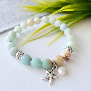 May include: A light green and white beaded bracelet with a silver starfish charm and a white pearl. The bracelet is on a white surface with green leaves in the background.
