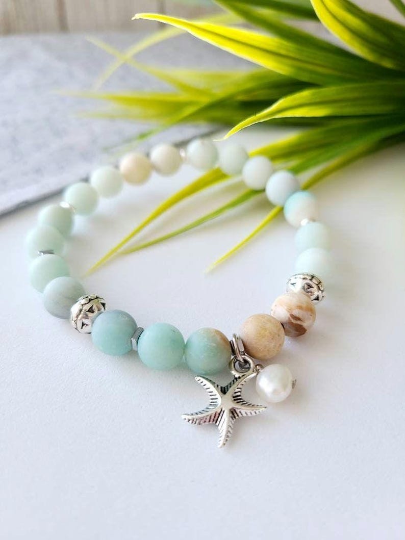 May include: A light green and white beaded bracelet with a silver starfish charm and a white pearl. The bracelet is on a white surface with green leaves in the background.