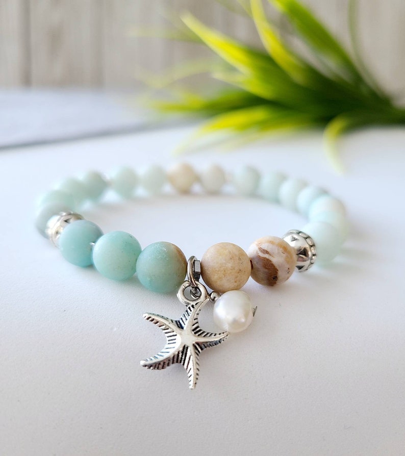 May include: A light blue and beige beaded bracelet with a silver starfish charm and a pearl. The bracelet is on a white surface.
