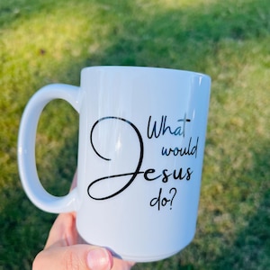 Puede incluir: Taza de cerámica blanca con un asa grande. La taza presenta el texto "Jesus" en una escritura estilizada, con las palabras "What would do?" en una fuente más pequeña y sencilla. La taza se sostiene sobre un fondo de hierba verde.