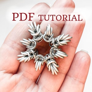 May include: A silver-colored, handmade snowflake ornament made of wire. The ornament is held in a hand and the text "PDF TUTORIAL" is displayed in burgundy letters above the ornament.