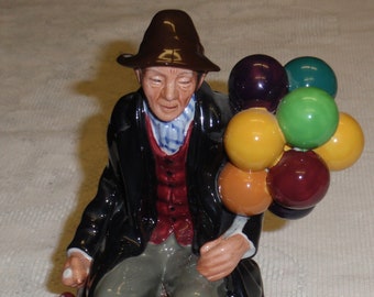 May include: A ceramic figurine of a man wearing a brown hat and a black coat, sitting on a wooden box and holding a bunch of colorful balloons.